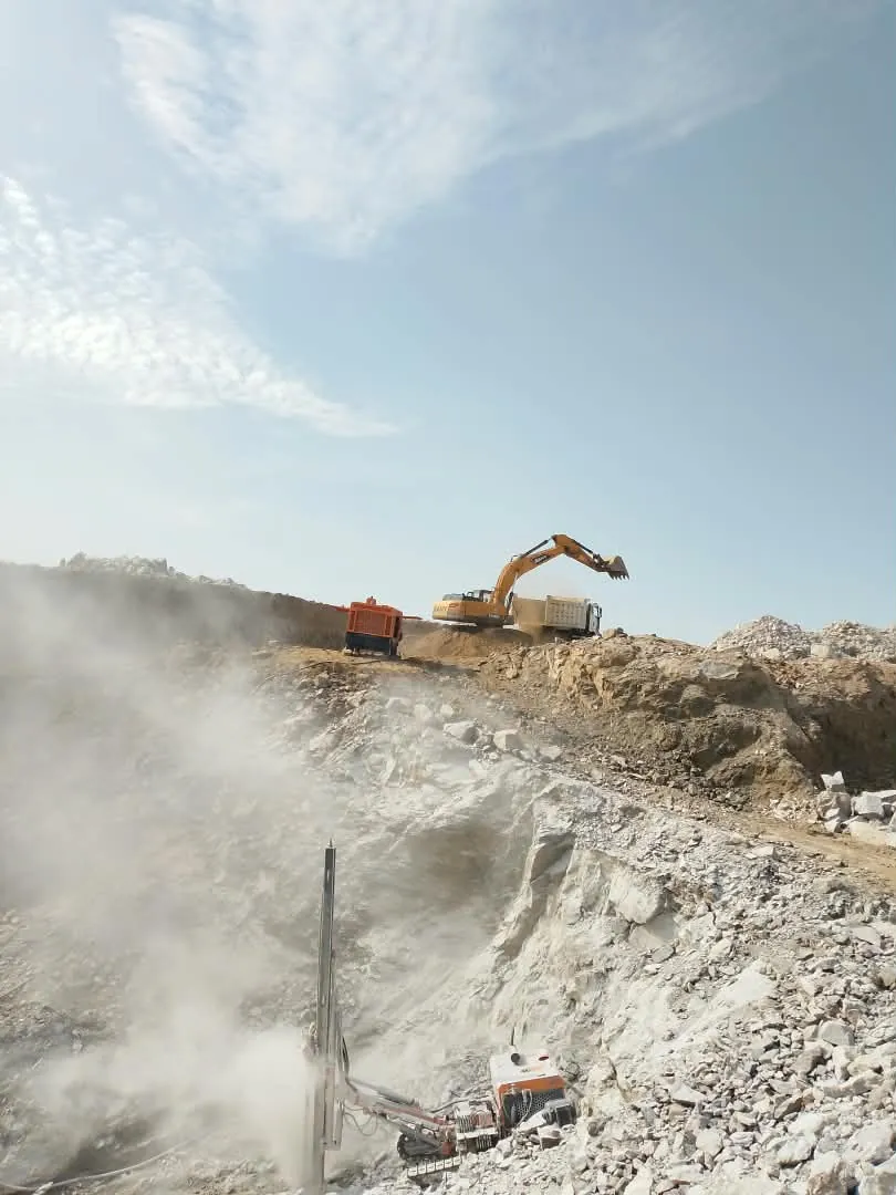 Drill rig in the foreground while an excavator loads a haul truck on the ridge above